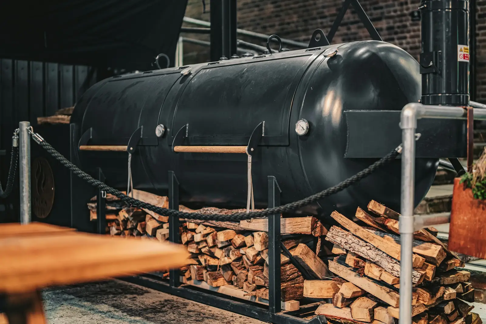 A black offset smoker built from an old tank, beside a stack of split wood